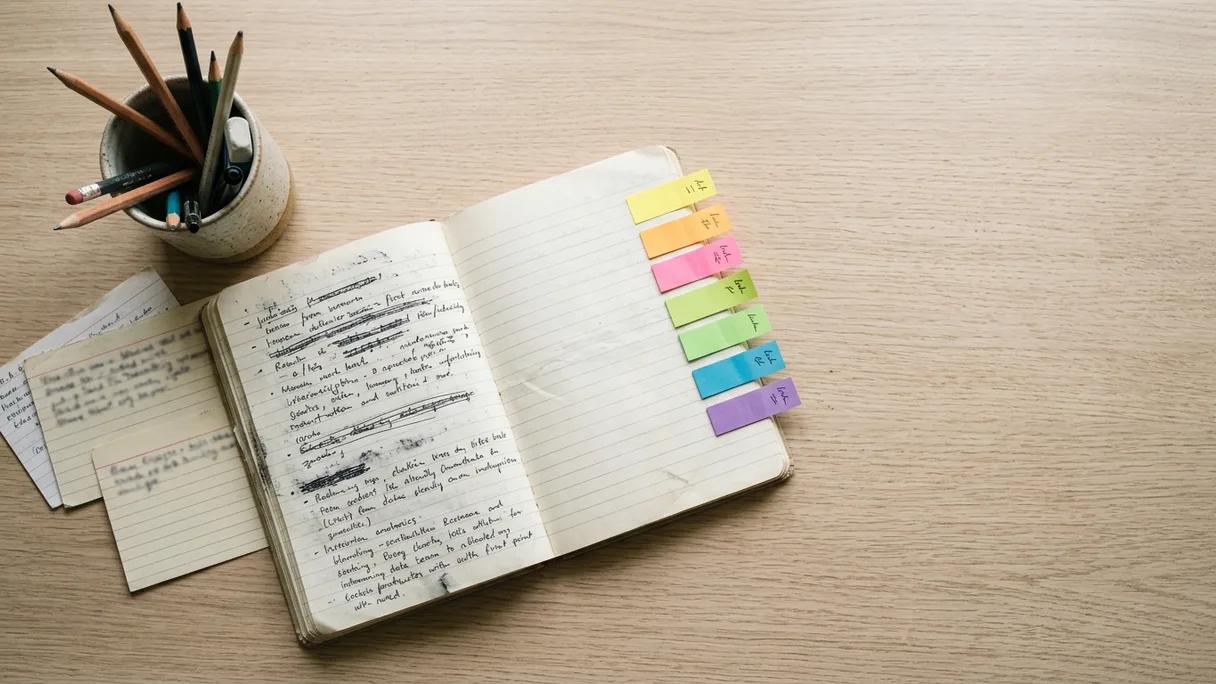 Overhead view of an open notebook with sticky notes and index cards on a sunlit oak desk.