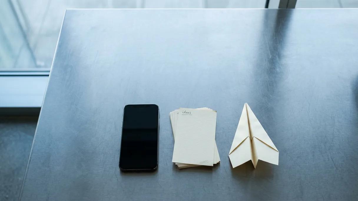 A phone face-down on a steel desk next to note cards and a folded paper airplane in cool light.